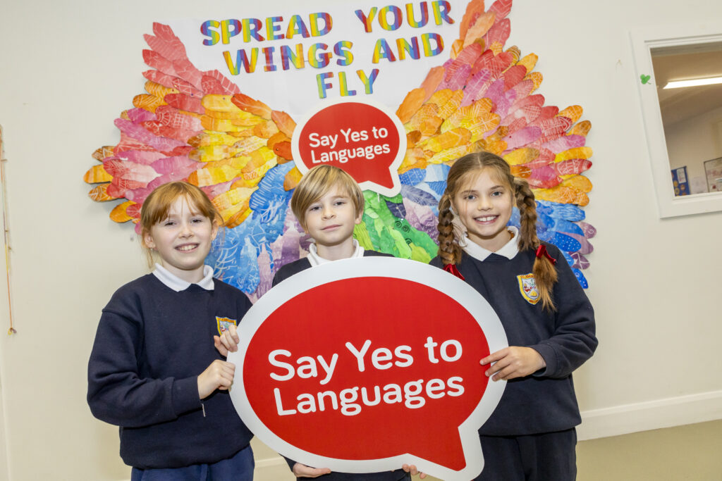Three primary pupils holding a Say Yes to Languages photo prop. They are smiling and standing in front of colourful angel wings.