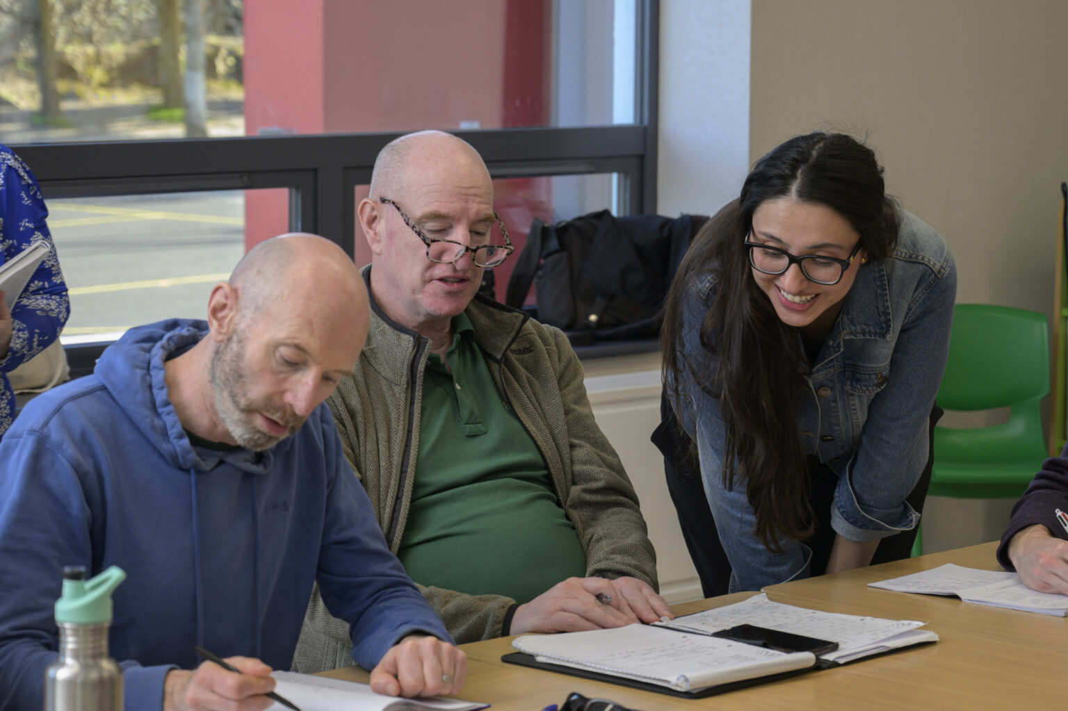 Two men sitting at a desk, looking down at their notebooks. Teacher leaning down to look at their work.